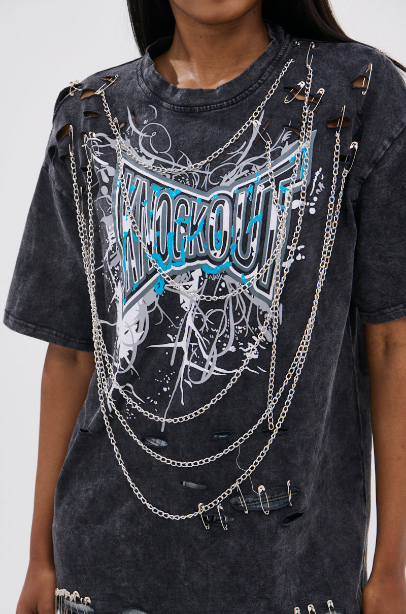 Wearing the KNOCK YOURSELF OUT DISTRESSED TEE, a black acid-washed top with "KNOCKOUT" in blue and white, styled with silver chains and safety pins. Their face is partly out of frame, creating an edgy, grunge-inspired look.