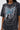 Wearing the KNOCK YOURSELF OUT DISTRESSED TEE, a black acid-washed top with "KNOCKOUT" in blue and white, styled with silver chains and safety pins. Their face is partly out of frame, creating an edgy, grunge-inspired look.