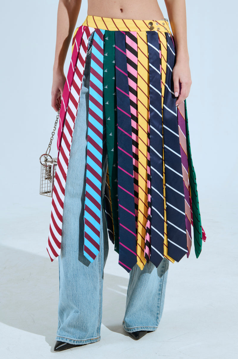 A person in jeans and a Tie Me Down Midi Skirt adorned with colorful tie patterns, metallic snap closures, and featuring stripes, polka dots, and paisley designs. They carry a small birdcage-shaped purse.