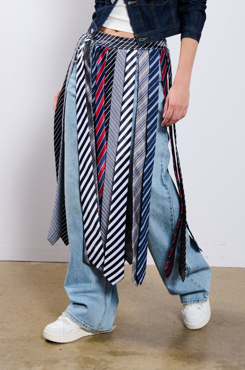 A person wearing wide-leg blue jeans, white sneakers, and a denim jacket. They are sporting the TIE ME DOWN MIDI SKIRT which features numerous men’s neckties in different striped patterns attached to their waist, creating a skirt-like look with a jagged hemline. The background is a plain white wall with a beige floor.