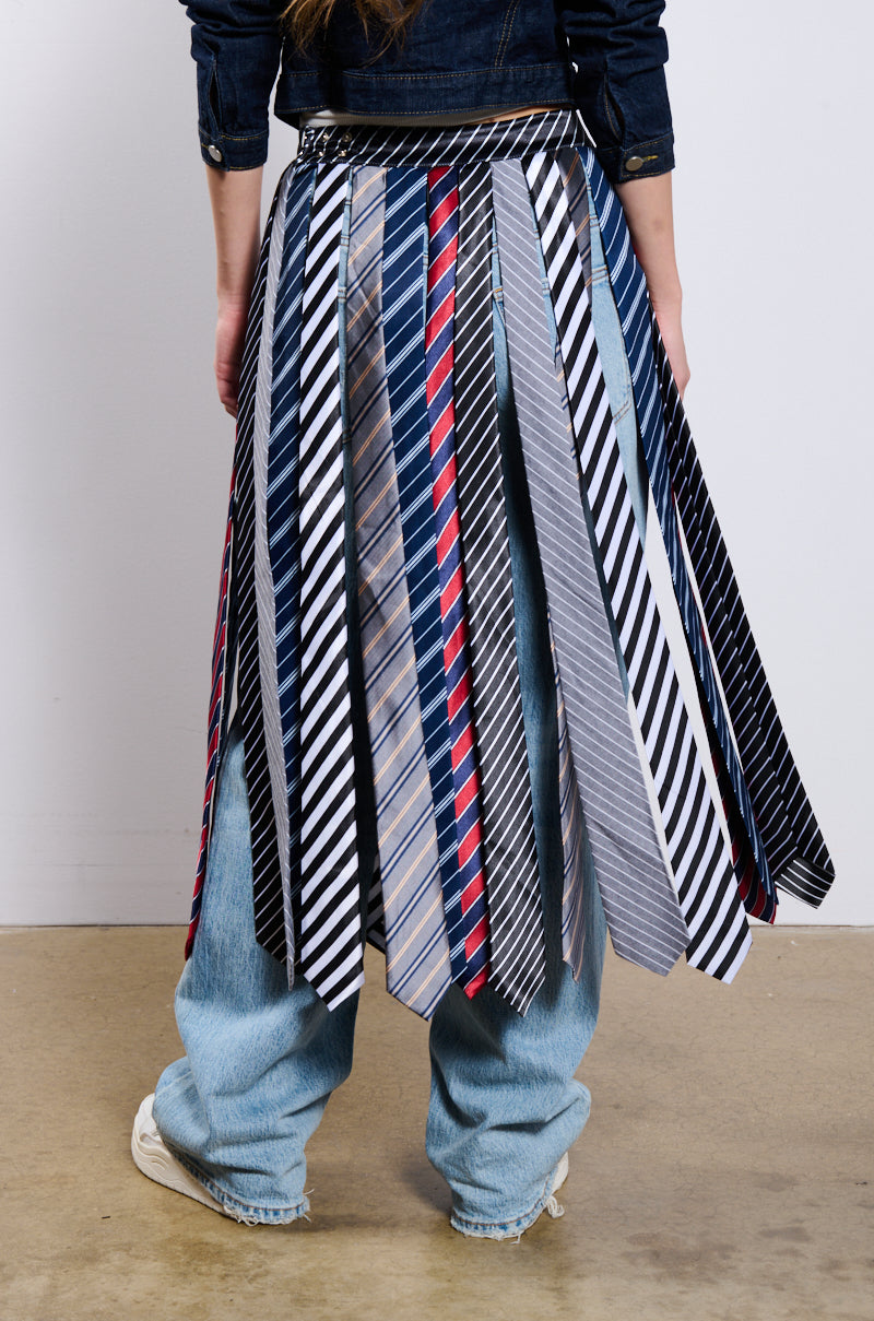 A person wearing the TIE ME DOWN MIDI SKIRT, made of variously patterned neckties with a jagged hemline, stands with their back to the camera. They are also wearing a denim jacket and light blue jeans. The skirt displays a mix of striped neckties, checks, and solid colored ties.