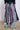 A person wearing the TIE ME DOWN MIDI SKIRT, made of variously patterned neckties with a jagged hemline, stands with their back to the camera. They are also wearing a denim jacket and light blue jeans. The skirt displays a mix of striped neckties, checks, and solid colored ties.