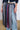 A person is wearing the TIE ME DOWN MIDI SKIRT, created entirely from various colored and patterned neckties, and featuring a jagged hemline. The skirt is paired with light blue jeans and a dark denim jacket against a plain white wall and brown floor background.