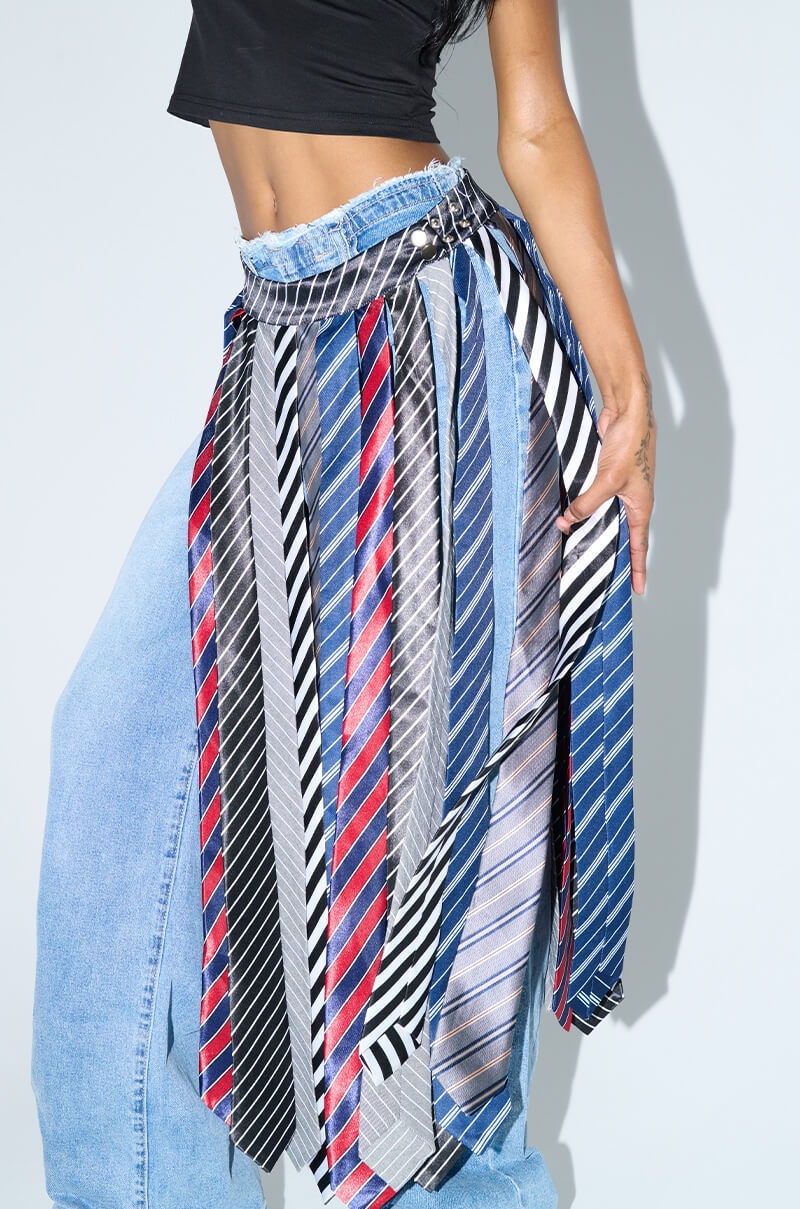 A person wearing a black crop top and blue jeans showcases the "TIE ME DOWN MIDI SKIRT," featuring a belt made from striped neckties draped around their waist. The ties display various patterns and colors, adding a unique and creative flair to the outfit.