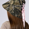 A person with long brown hair is wearing the "LACE ME UP CAMO BUCKET HAT," which features pink satin lace-up ribbons on the back. The plain, light-colored background emphasizes the design and intricate details of the hat.