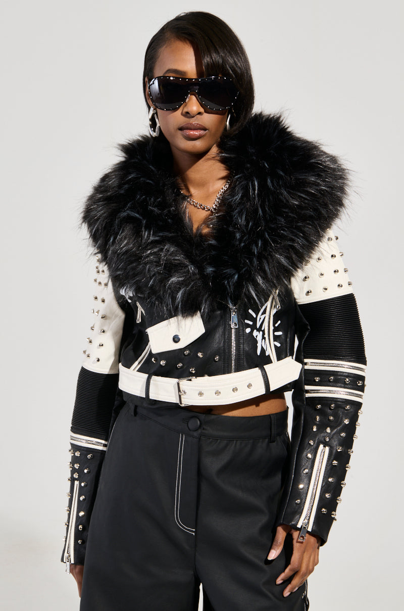 Dressed in the FROM THE BRONX FUR TRIM MOTO jacket, a fashionable black and white faux leather piece with a fur collar and studded accents, the person pairs it with sleek black pants. Adorned with large dark sunglasses and hoop earrings, they exude confidence against a neutral backdrop.