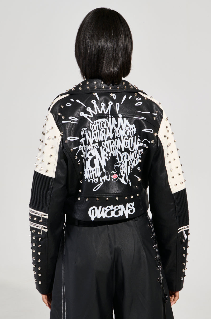 A person with medium-length hair wears the FROM THE BRONX FUR TRIM MOTO jacket. This black and white streetwear piece is decorated with graffiti-style writing and embellishments, highlighting the word "QUEENS" at the bottom. Complete with metal studs, abstract designs, and an edgy fur trim, this faux leather jacket captures the ultimate moto vibe.