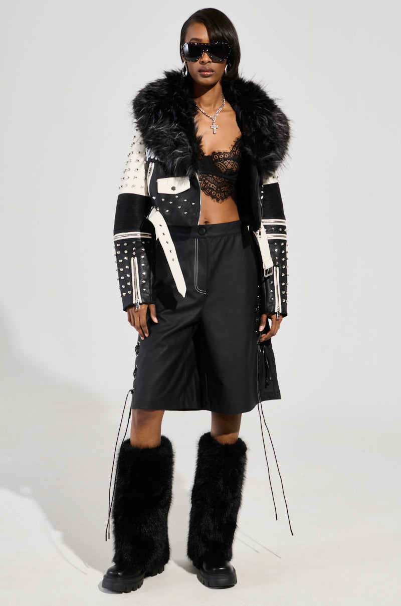 Wearing the FROM THE BRONX FUR TRIM MOTO jacket with confidence, a person shows off a stylish lace top, knee-length leather shorts, black furry boots, and sunglasses. Layered necklaces complete their streetwear ensemble.