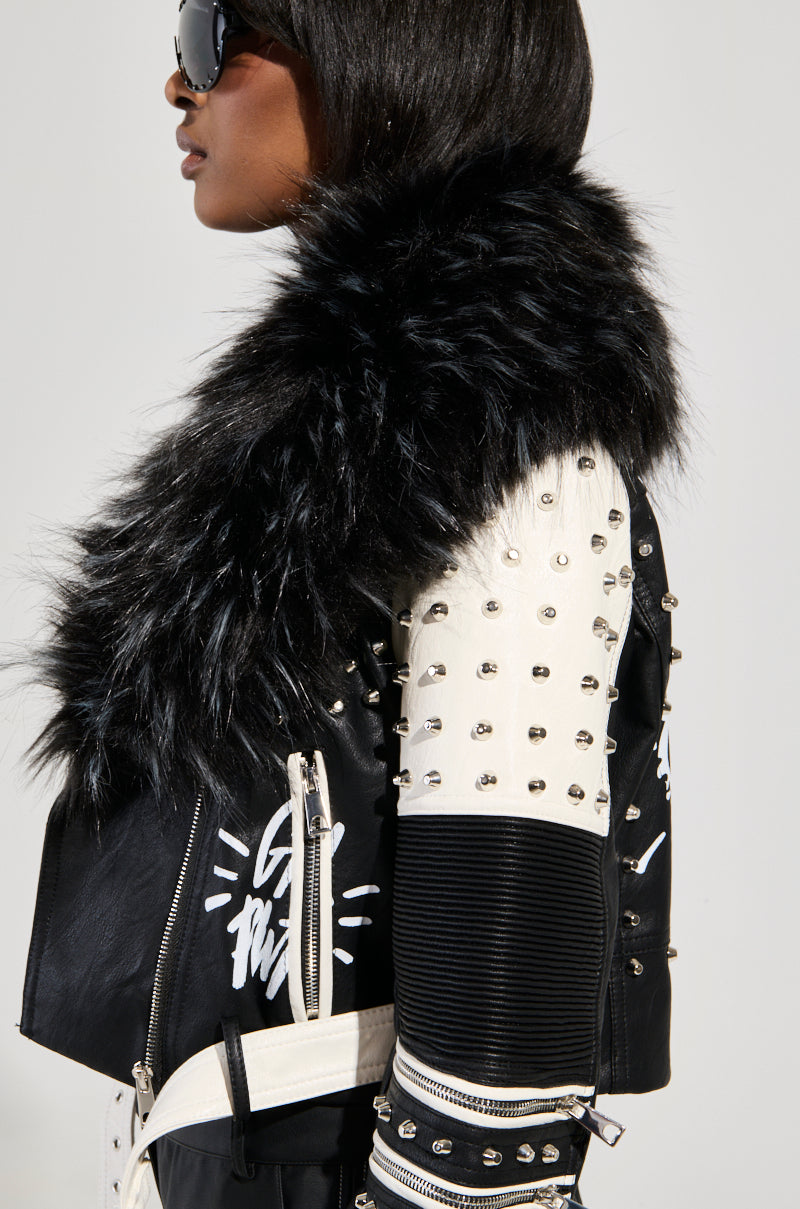 A person wearing sunglasses and the FROM THE BRONX FUR TRIM MOTO, a striking jacket with a studded black and white design. This faux leather streetwear piece boasts metallic studs and zippered sleeve details, delivering an effortlessly punk-inspired look.