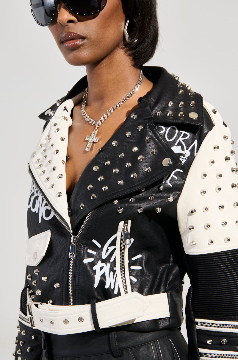 A person sports the FROM THE BRONX FUR TRIM MOTO jacket, a black and white streetwear piece embellished with graffiti-style writing. This studded design is accessorized with a silver cross necklace, large sunglasses, and showcases a distinctive belt with mixed textures, embodying edgy urban style.
