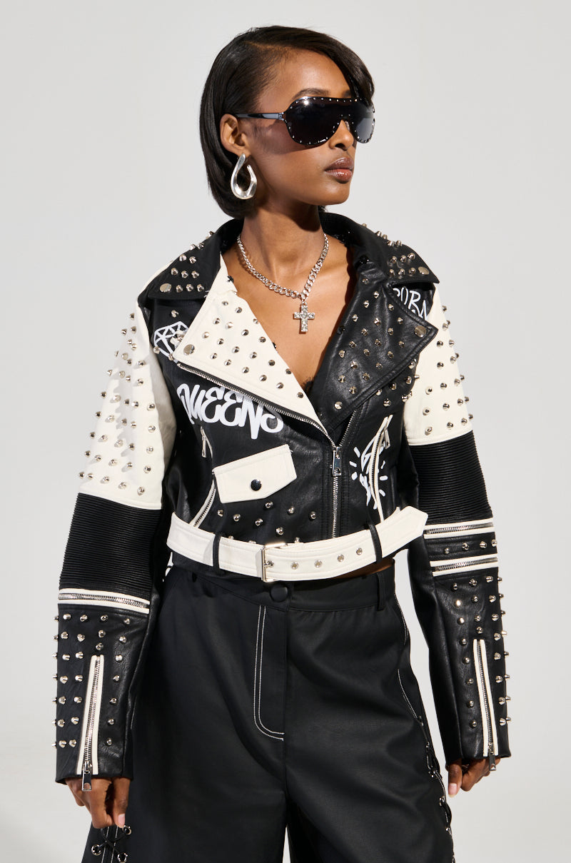 A person confidently stands with hands in pockets against a plain gray background, wearing the FROM THE BRONX FUR TRIM MOTO jacket, which features black and white faux leather adorned with studs and silver embellishments. They accessorize with black sunglasses and hoop earrings, while sporting a short hairstyle.