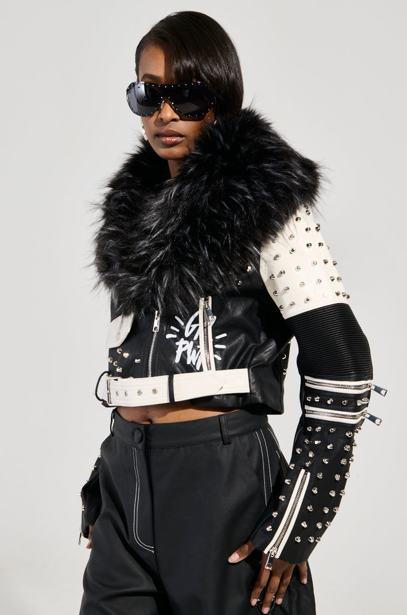 A person wearing stylish black sunglasses, black pants, and the FROM THE BRONX FUR TRIM MOTO jacket featuring a studded design and zippers. The jacket showcases graphic elements that embody streetwear chic. The person strikes a confident pose against a neutral background.