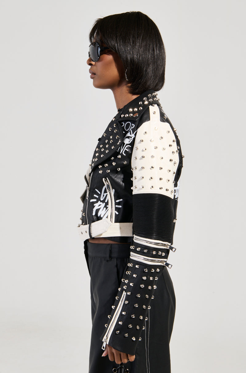 Wearing the FROM THE BRONX FUR TRIM MOTO jacket, a person stands in profile showcasing its studded black and white streetwear aesthetic with text designs. They complement their bold style with short hair and dark sunglasses against a plain, light-colored background that highlights their edgy look.