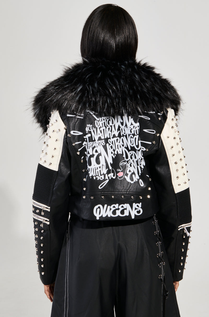 A person wearing the FROM THE BRONX FUR TRIM MOTO jacket, featuring black and white faux leather with fur trim, metallic studs, and graffiti-style graphics. With long dark hair cascading down, they stand confidently against a plain gray background, embodying the edgy spirit of streetwear fashion.