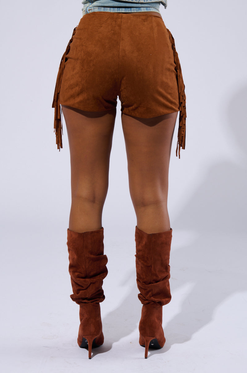 A person stands with their back to the camera, exuding a Coachella Revival vibe in THE RODEO SHOW SHORT IN BROWN SUEDE, paired with a denim top and slouchy brown knee-high suede boots with heels. The background is plain light gray.