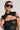 A person wearing the TRUE TRUE PU COLLAR is posing against a neutral background. They have their hair slicked back, are wearing large black sunglasses, and have long dangling earrings. The black leather top pairs seamlessly with the sleeveless silhouette enhanced by the black studded harness draped over their shoulders, which they flaunt with faux leather accessories.