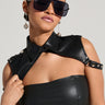 A person wearing the TRUE TRUE PU COLLAR is posing against a neutral background. They have their hair slicked back, are wearing large black sunglasses, and have long dangling earrings. The black leather top pairs seamlessly with the sleeveless silhouette enhanced by the black studded harness draped over their shoulders, which they flaunt with faux leather accessories.