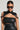 A person wears a black, sleeveless silhouette top paired with a black leather harness embellished with metallic studs and the standout TRUE TRUE PU COLLAR. They complete the look with large, dark sunglasses and dangling spherical earrings, while their hair is styled sleekly pulled back.