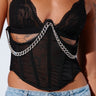 A close-up image of a person wearing a black lace bralette and the NOT BASIC CHAIN DETAIL CORSET BELT adorned with a silver chain across the front. The person is also wearing light blue denim jeans with embellishments and layered silver necklaces, one featuring a cross pendant.