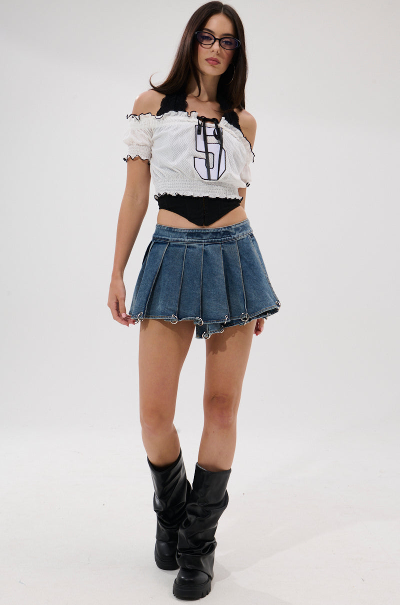 A woman in an off-the-shoulder white crop top with black trim, the TOO MANY NIGHTS LIKE THIS DENIM MINI SKIRT, black knee-high boots, and glasses stands against a plain white background. 4