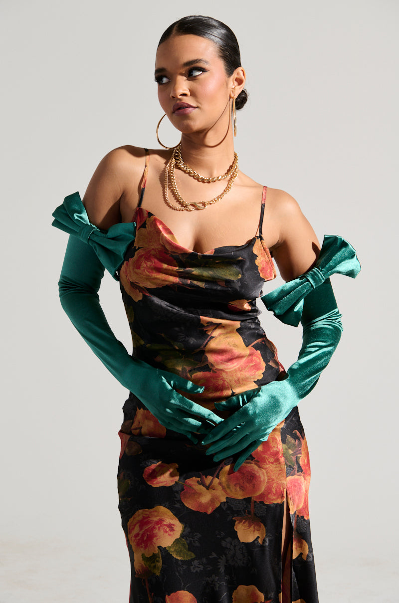 A person poses confidently in a floral dress with a cowl neckline, adorned with luxurious accessories. They wear long, GIVING RICH SATIN GLOVES in teal and gold hoop earrings, their hair styled back. The background is plain and light-colored. 0