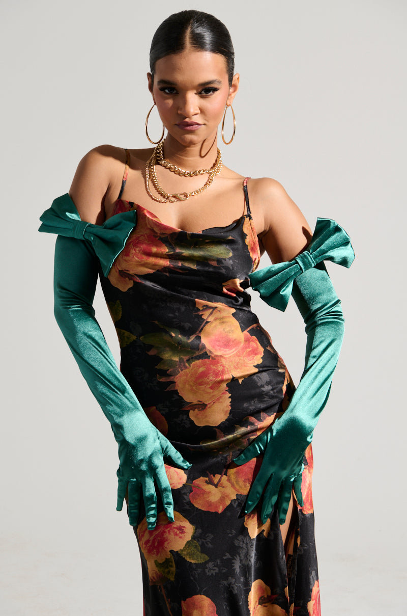 A person confidently poses in a floral dress featuring a slit, accompanied by the GIVING RICH SATIN GLOVE adorned with oversized bows. Their look is completed with hoop earrings, a chunky necklace, and smoothed-back hair, all set against a plain background for an added touch of style. 1