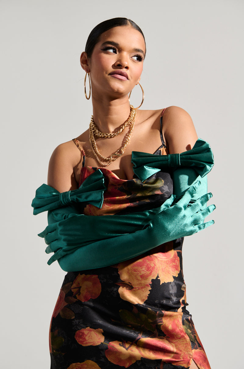 A woman poses confidently in a floral dress, showcasing the GIVING RICH satin gloves adorned with oversized bows. She accessorizes with gold hoop earrings and layered necklaces, while the plain background highlights her striking style, emphasizing every luxurious accessory she wears. 2