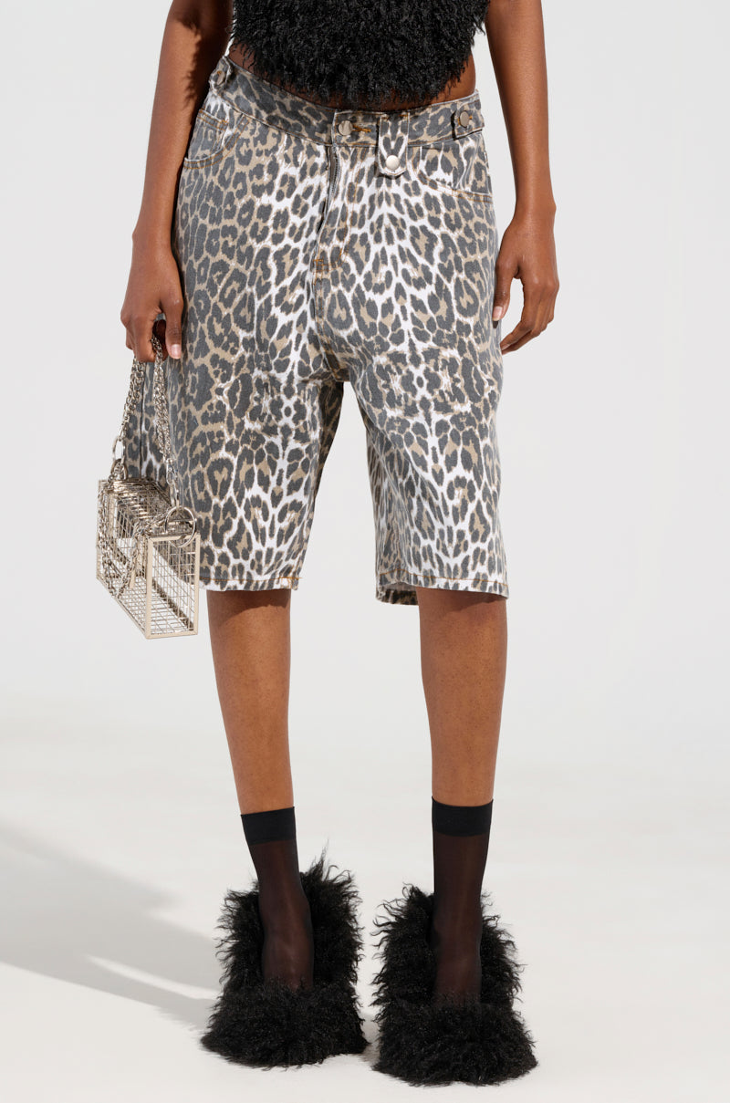 A person wearing RUN BABY RUN ANIMAL PRINT BERMUDA SHORTS, teamed with a fuzzy black top and black fluffy shoes, holds a small, clear box-shaped bag against a plain, light-colored background.