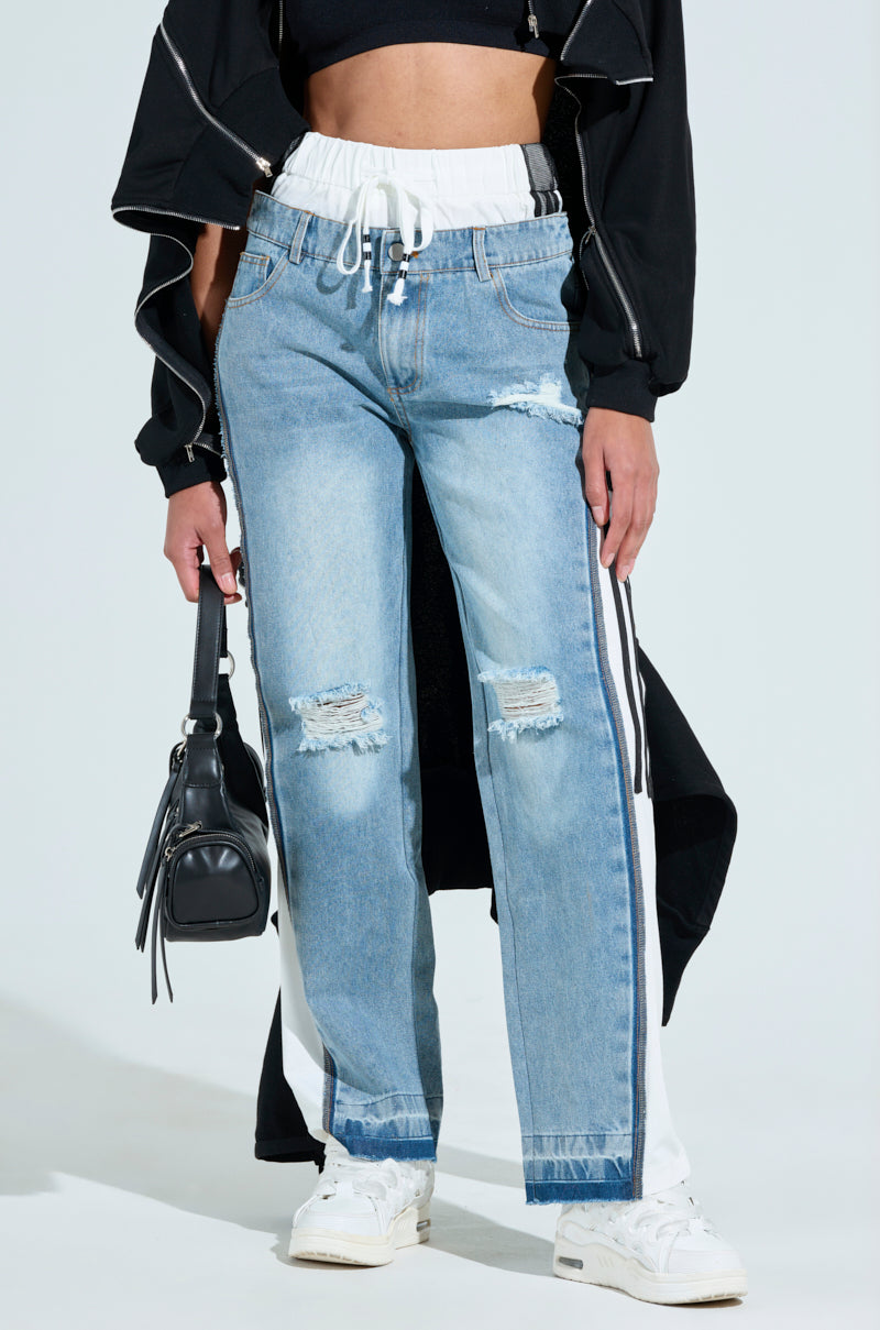 A person in GET WITH IT DENIM TRACK PANT, with side stripes and distressed patchwork at the ankles. They wear a black cropped jacket, white sneakers, and hold a small black handbag. The jeans feature blue fabric, five-pocket design, and a white drawstring waistband.