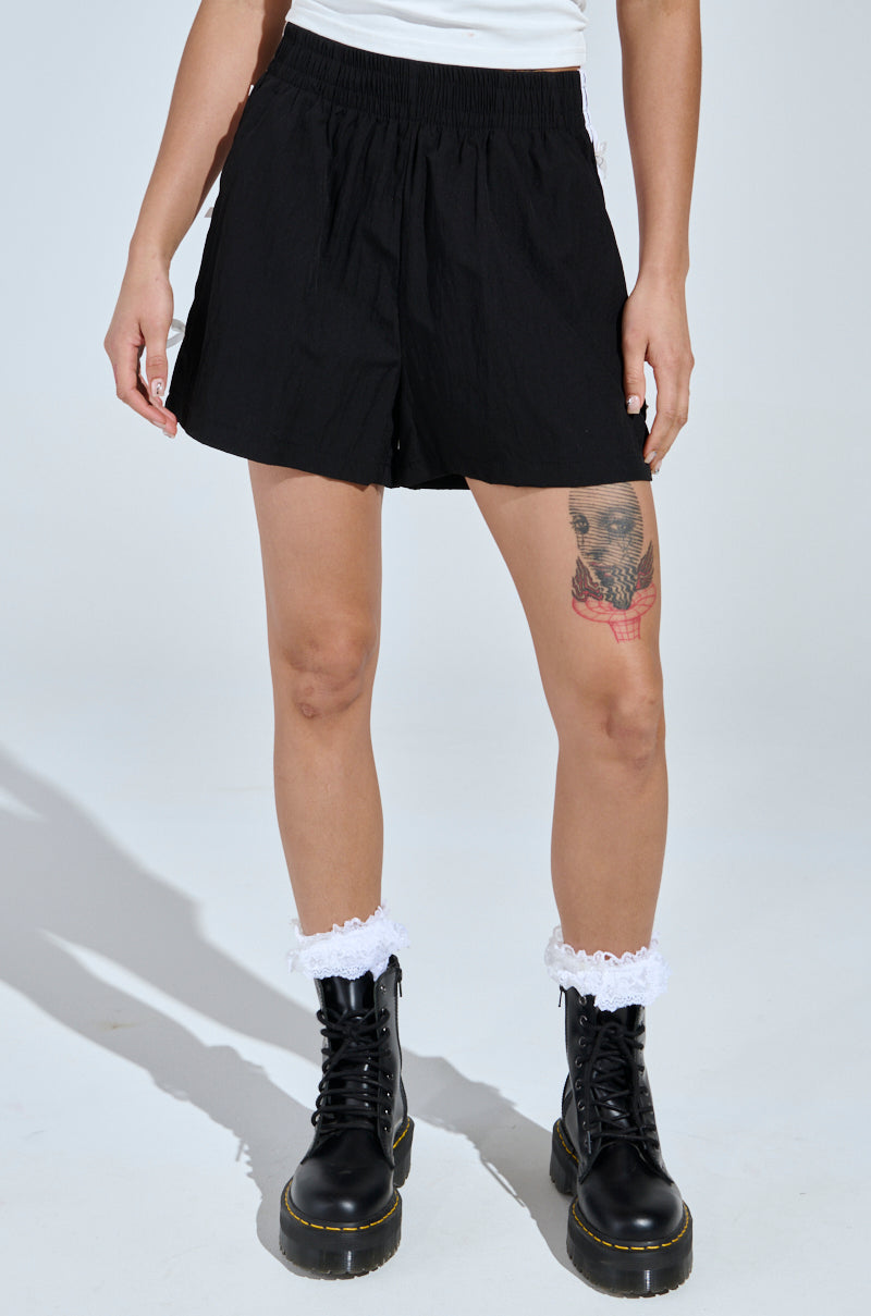 Dressed stylishly in a white top, BELLA BABY SHORT WITH BOW, and black combat boots paired with white lace socks, they showcase a red skull tattoo on their left knee against a minimalist white backdrop, embodying the sporty-chic vibe.