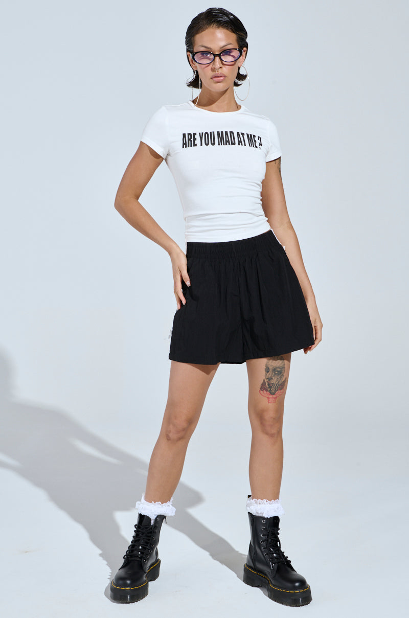 A person wears glasses and a white t-shirt reading "ARE YOU MAD AT ME?" paired with the BELLA BABY SHORT WITH BOW in black, combining sporty-chic flair with black boots and white socks, confidently displaying their leg tattoo.