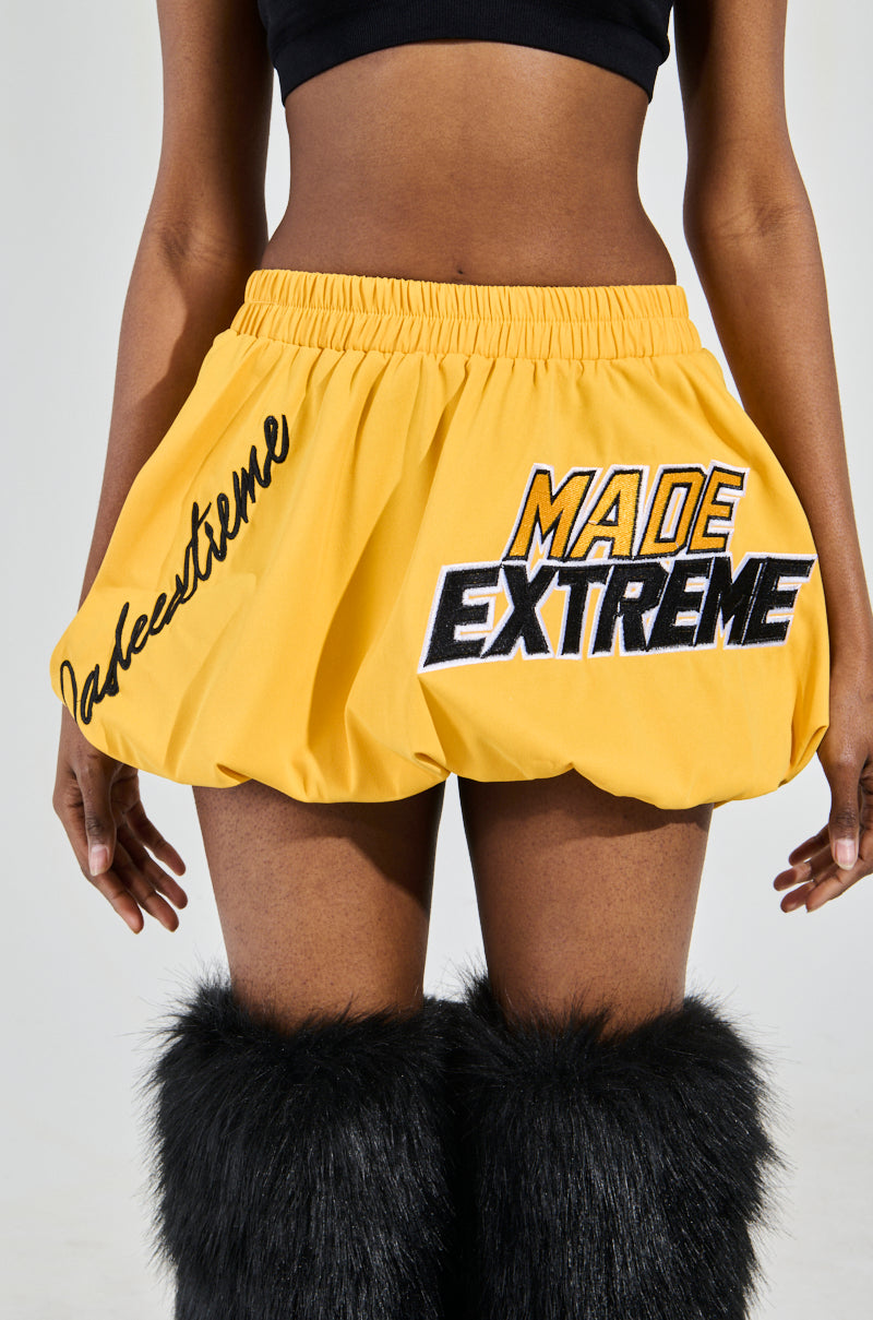 Someone wears the yellow MADE EXTREME BUBBLE SKIRT, featuring racing-inspired "MADE EXTREME" and "tasteetreme" text. They pair it with a black crop top and black fluffy leg warmers, standing against a plain background.
