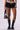 A person is wearing the Overthinking Is Great Mini Skirt in Black, featuring black tiered ruffle detailing and an adjustable grey drawstring closure. They pair it with black knee-high boots with thick soles and hold a small black handbag. The plain white background keeps the focus on the lower half of their body.