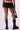 A person is shown from the back wearing a black OVERTHINKING IS GREAT MINI SKIRT IN BLACK with tiered ruffle detailing, black chunky knee-high boots, and holding a black handbag with a shoulder strap. The background is a plain off-white.