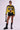 A woman stands confidently in a bright yellow and black racing jacket adorned with logos, a cropped gray top, and an Overthinking Is Great Mini Skirt In Black. She accessorizes with black sunglasses, chunky black boots, and a small black handbag, striking a fashionable pose.