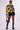 A person poses confidently wearing a yellow and black racing-themed jacket, a grey crop top, and the Overthinking Is Great Mini Skirt in Black with tiered ruffle detailing. They finish the look with black knee-high boots, black sunglasses, and a matching handbag.