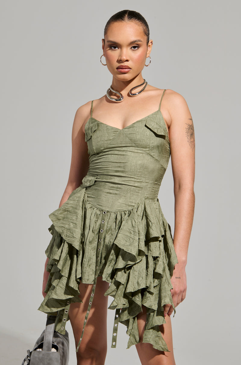 A person stands confidently in a green, textured CANT COMPARE CHIFFON MINI DRESS by AKIRA Label, featuring ruffled layers and thin straps. They accessorize with hoop earrings, a bold necklace, and carry a gray bag, all set against a plain light gray backdrop.