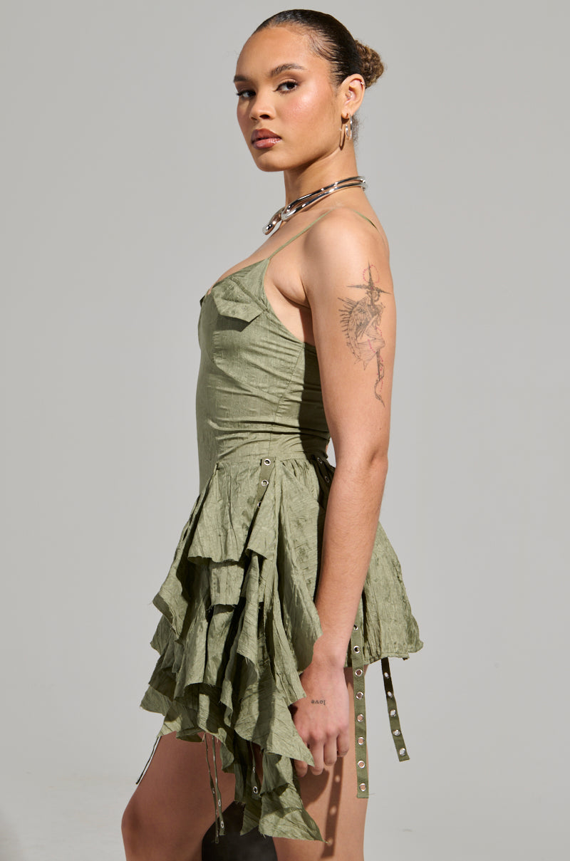 A person wearing the CANT COMPARE CHIFFON MINI DRESS in green poses confidently against a neutral background. Their hair is styled in a neat bun, and they have a tattoo on their arm. A necklace and earrings enhance their stylish look, highlighting the dress's elegant ruffled skirt.