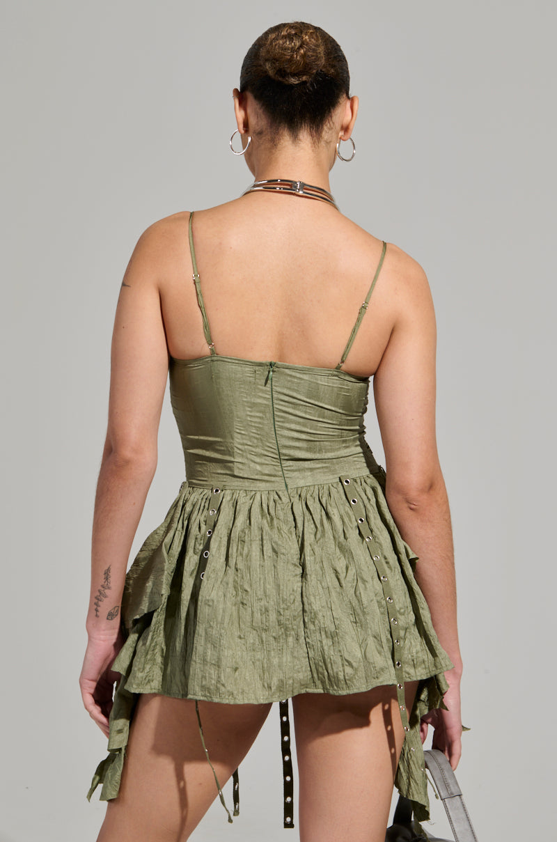 Rear view of a person wearing the CANT COMPARE CHIFFON MINI DRESS in olive green, featuring thin straps and a pleated skirt. They have short hair styled in a bun and complement the look with hoop earrings and a choker. A tattoo on their left arm provides an edgy contrast to the elegant AKIRA Label ensemble.
