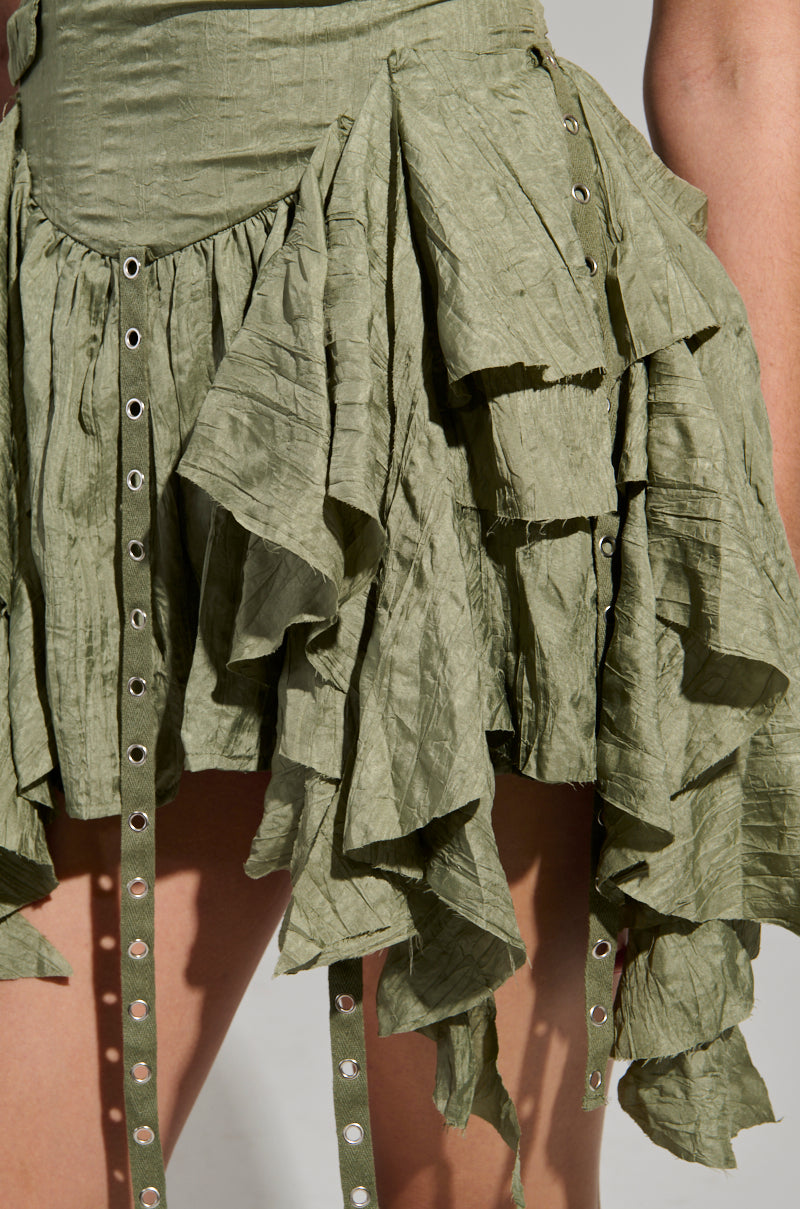 A close-up of a textured olive green CANT COMPARE CHIFFON MINI DRESS by AKIRA Label, featuring layered ruffles and metallic eyelet details. The fabric has a crinkled appearance, with decorative straps adorned with rivets hanging down.