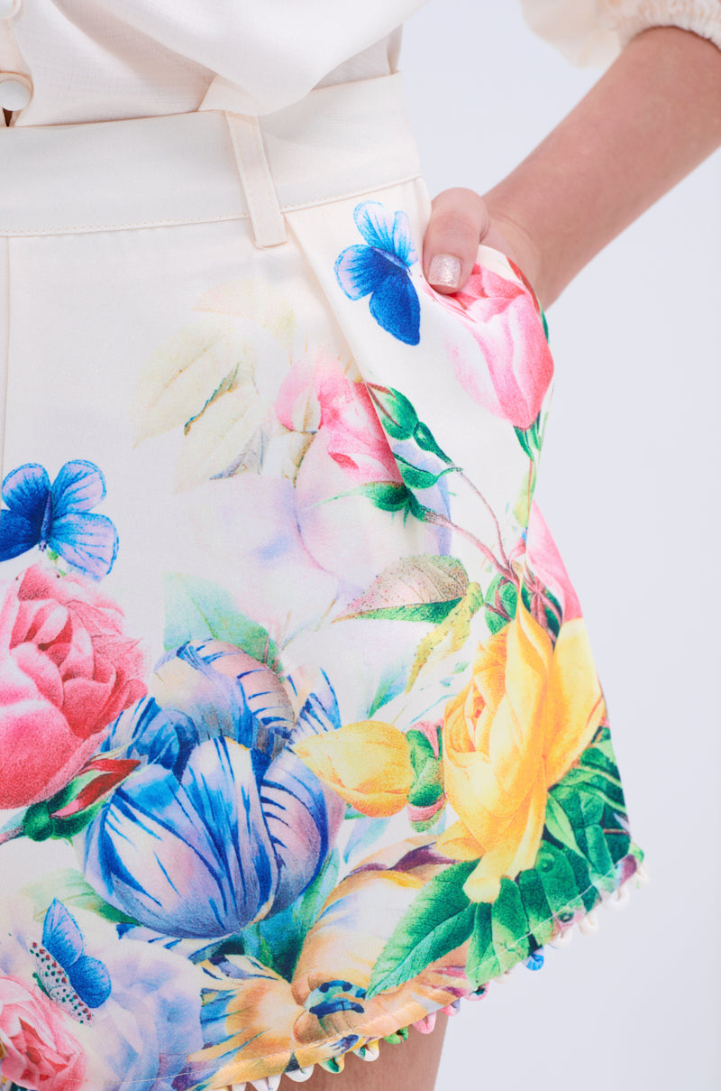 Close-up of a person wearing the ROSE FLORAL SHORT, featuring a high-rise cream design with vibrant floral and butterfly prints in pink, yellow, blue, and green—perfect for garden parties. Hand rests casually in the pocket.