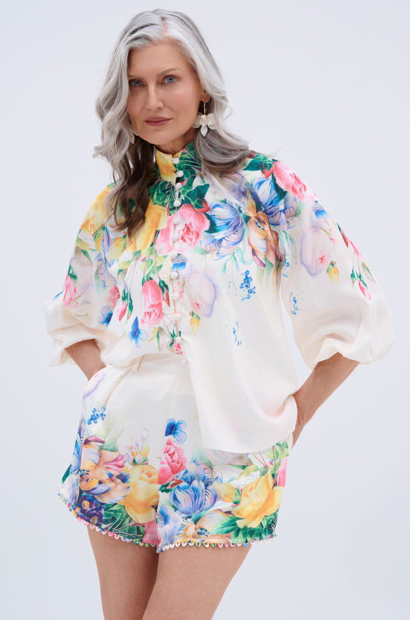 A confident woman with long, wavy gray hair smiles gently against a plain backdrop, wearing the ROSE FLORAL BUTTON DOWN and matching floral shorts—colorful flowers on white lending her look a fresh, botanical-inspired style.