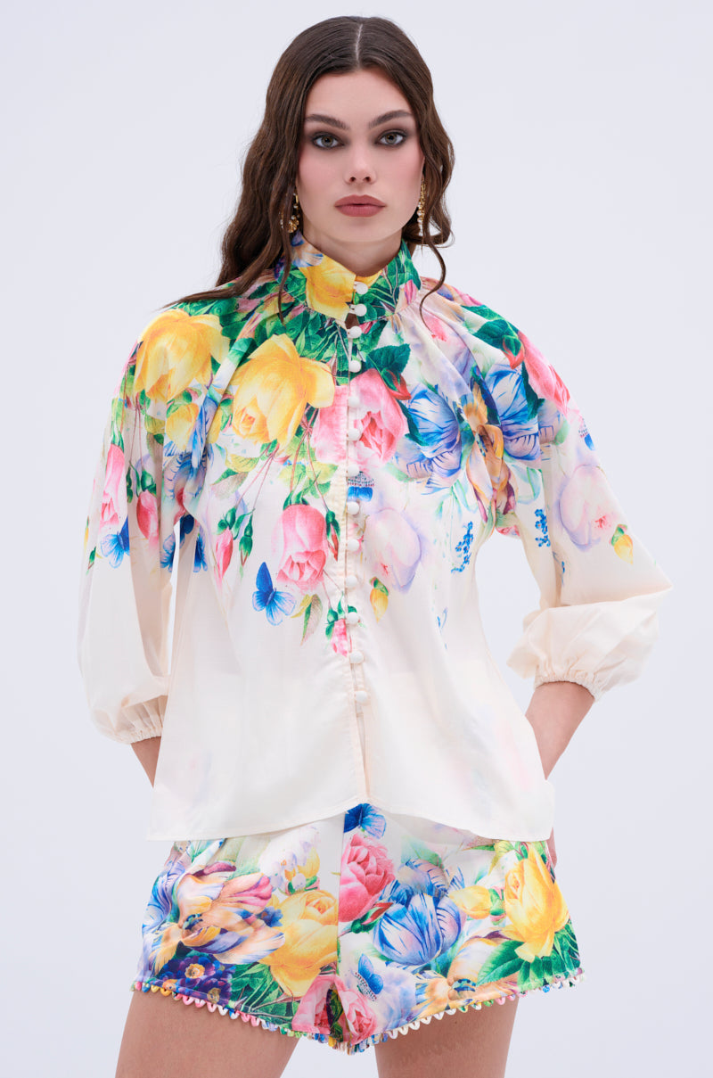 A woman wears the ROSE FLORAL BUTTON DOWN, a high-neck, long-sleeve blouse with colorful pink, yellow, blue, and green flowers. She pairs it with floral shorts and stands hands-in-pockets against a light background.
