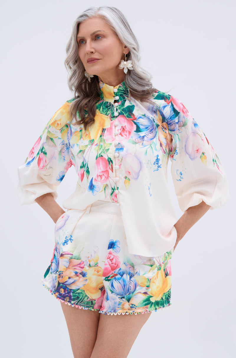 A smiling older woman with long gray hair wears the ROSE FLORAL BUTTON DOWN, featuring colorful flower prints. She confidently poses with her hands in her pockets.