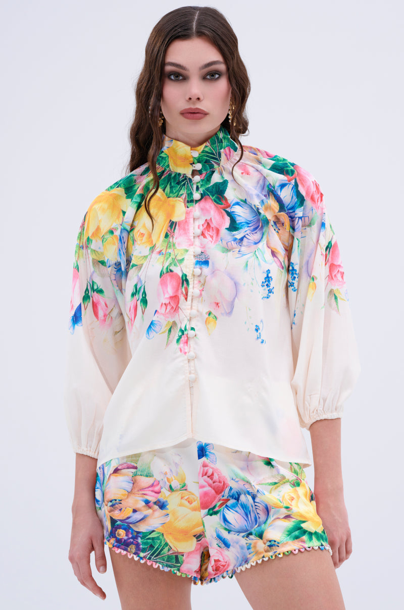 A woman with long wavy brown hair wears the ROSE FLORAL BUTTON DOWN, a cream blouse with a colorful floral print, high neck, and puffed sleeves, paired with matching floral shorts. She stands against a plain light background.