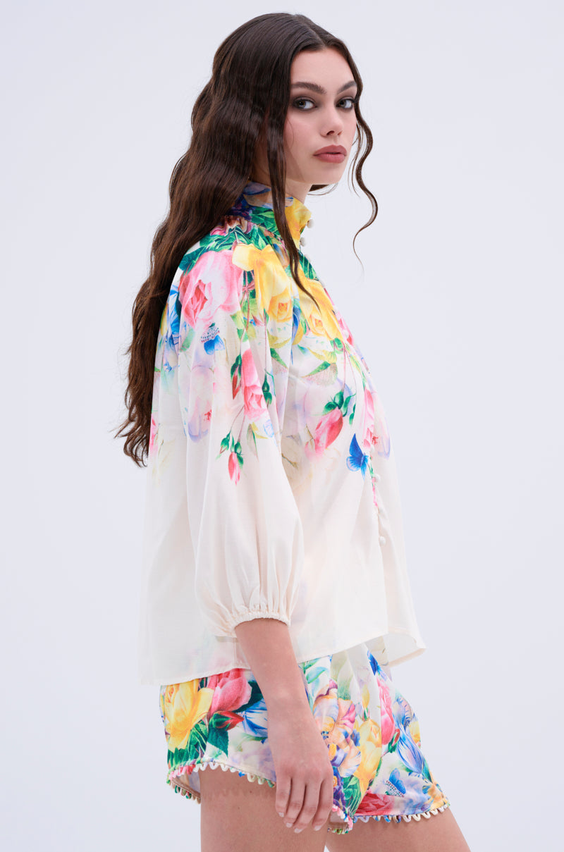 A woman with long wavy brown hair wears the ROSE FLORAL BUTTON DOWN and matching floral shorts with pompom trim, standing sideways against a light background.