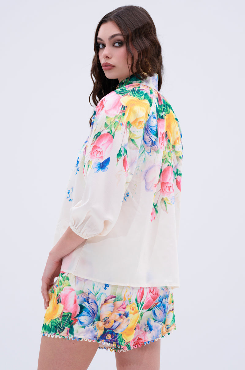 A woman with wavy brown hair wears the ROSE FLORAL BUTTON DOWN and matching cream floral shorts with vibrant pink, yellow, and blue flowers, standing against a plain light background.