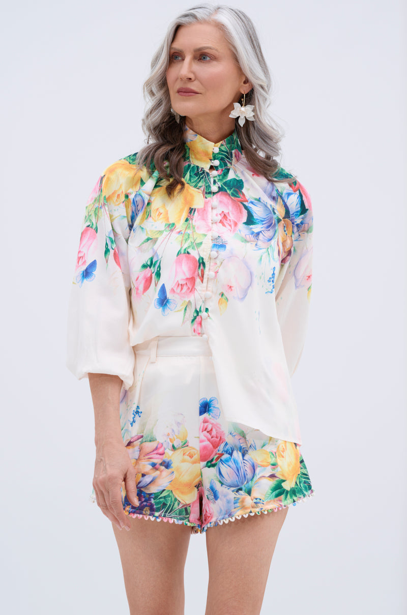 A woman with long gray hair wears the ROSE FLORAL BUTTON DOWN, featuring colorful pink, yellow, blue, and green flowers on a white background. She pairs it with floral shorts and smiles slightly while looking to the side.