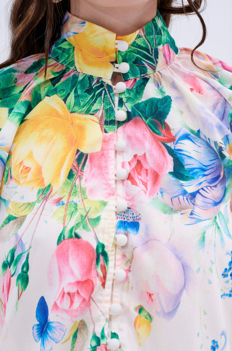 A close-up of the ROSE FLORAL BUTTON DOWN displays vibrant roses, lush greenery, and blue butterflies. It features a high collar and white fabric-covered buttons on the front.