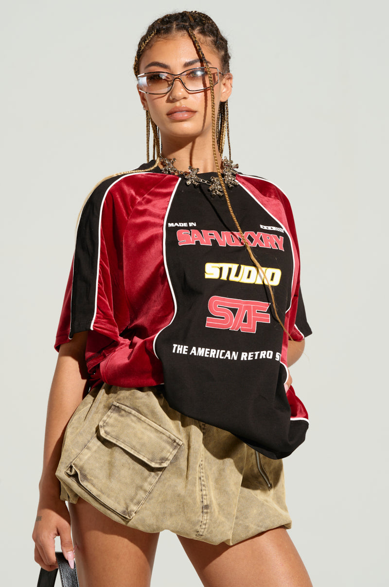 A person with long braided hair wearing sunglasses, a bold red and black oversized THE AMERICAN JERSEY with colorblock paneling, and beige cargo shorts poses confidently against a neutral background.